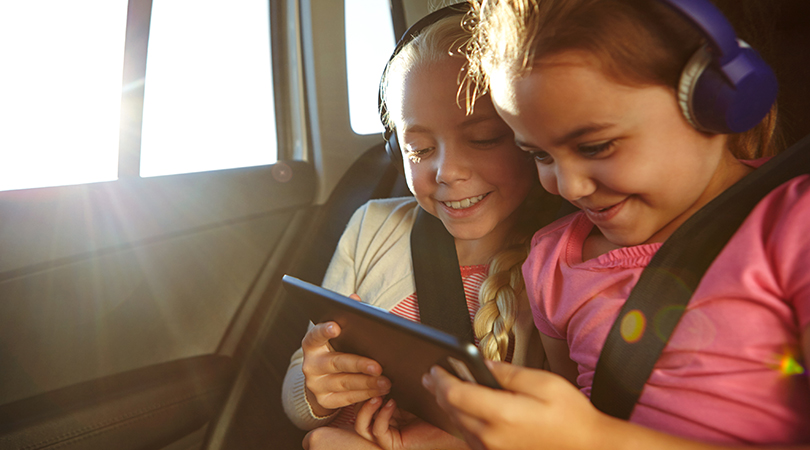 Two children with headphones sit in the car and look smiling at a tablet
