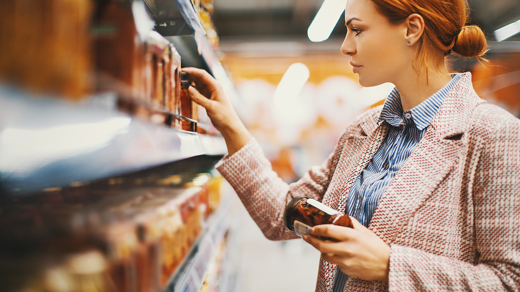 A woman taps on a smartphone in front of a supermarket shelf.
