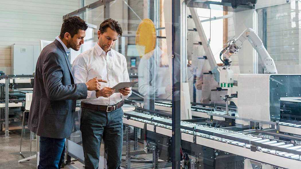 Two men stand in front of a production line and look together at a tablet
