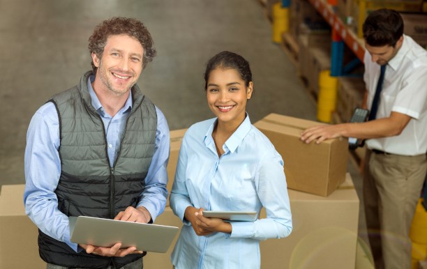 A man holding a tablet and a woman smile at the camera, in the background a warehouse worker scans a package.