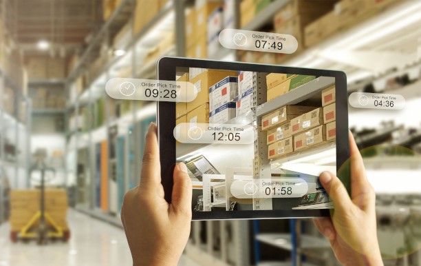 Two hands hold up a tablet in a warehouse, on and next to the screen are virtual boxes with information about the goods.