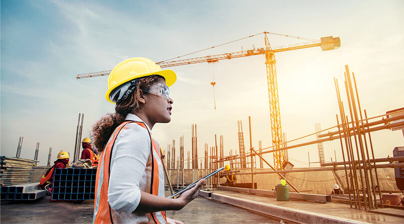 Woman with safety helmet, safety glasses, high-visibility vest and tablet looks at a construction site