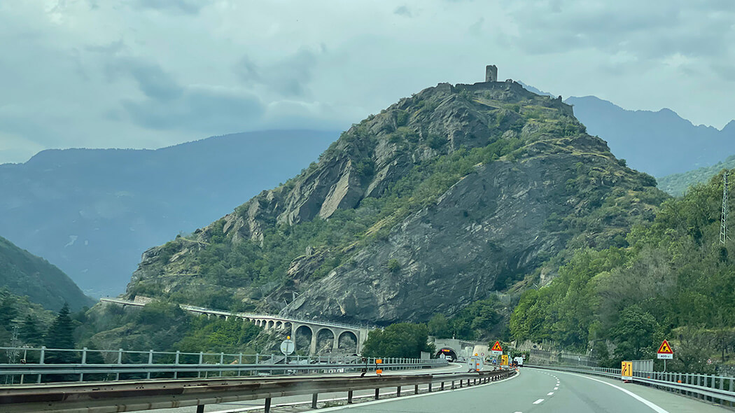 The autobahn toward Mont Blanc heads for a tunnel beneath a mountain topped by a ruined castle.