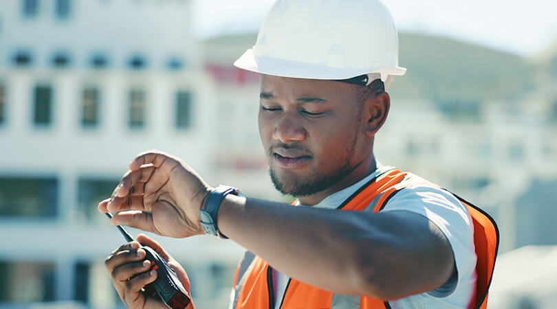 Mann in Schutzausrüstung für die Baustelle und Funkgerät in der Hand blickt auf seine Armbanduhr