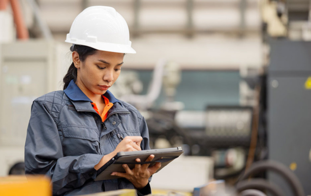 Frau mit Helm in Fabrikhalle schreibt auf ein Tablet