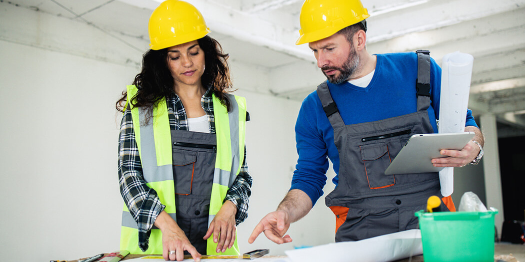 Man and woman wearing a yellow protection helmet and pointing at construction plans.