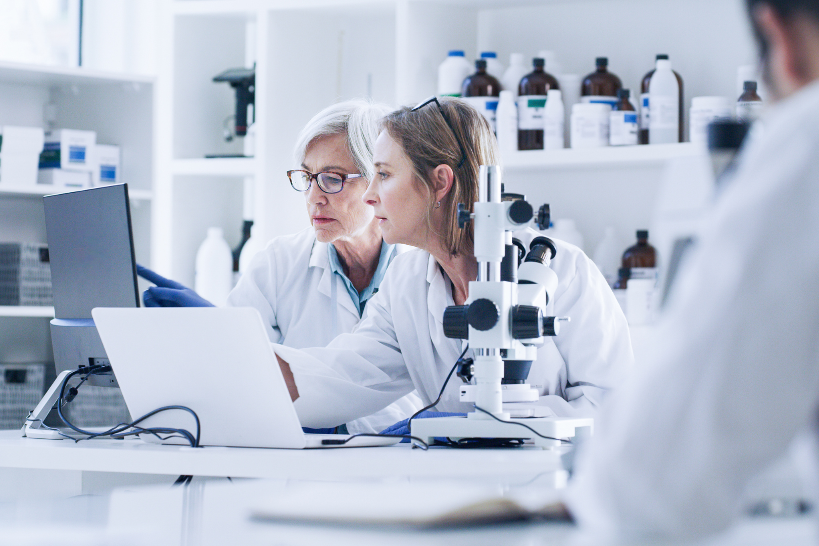 Two female scientists in lab coats are looking at a laptop together in a lab.