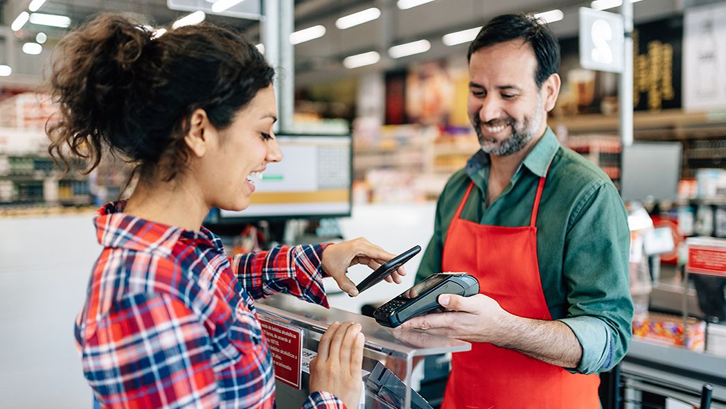 Frau bezahlt im Supermarkt kontaktlos mit dem Smartphone bei einem Mitarbeiter.