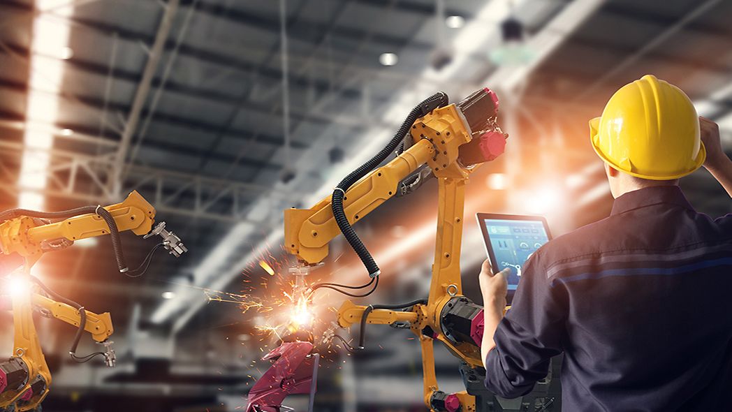 Mann mit Tablet in der Hand steht in einer Werkshalle vor Robotern.