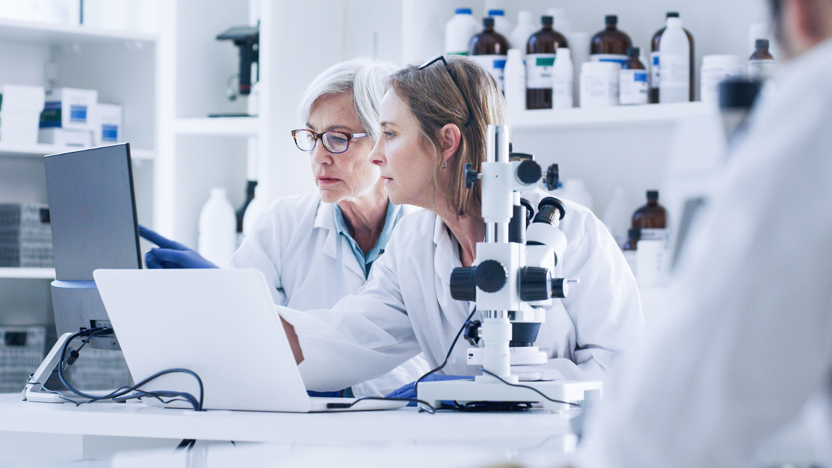Two female scientists in lab coats are looking at a laptop together in a lab.