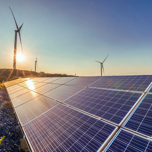 Solar modules in a field reflecting the sun, with wind turbines in the background.