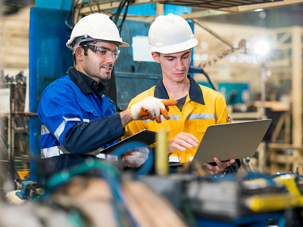 Two workers wearing hard hats check data on a laptop in a factory