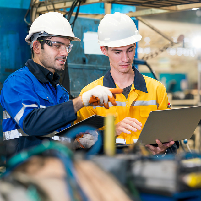 Two workers wearing hard hats check data on a laptop in a factory