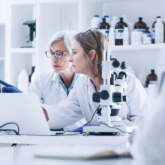 Two female scientists in lab coats look together at a laptop in a laboratory.