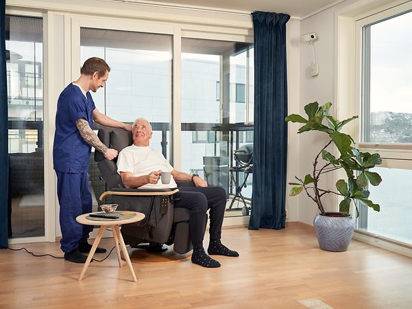 A carer is standing next to an elderly person sitting in a chair.