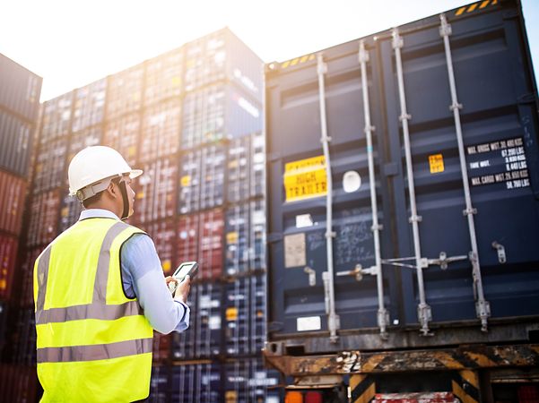 An employee in a high-visibility vest and helmet checks container data on his smartphone