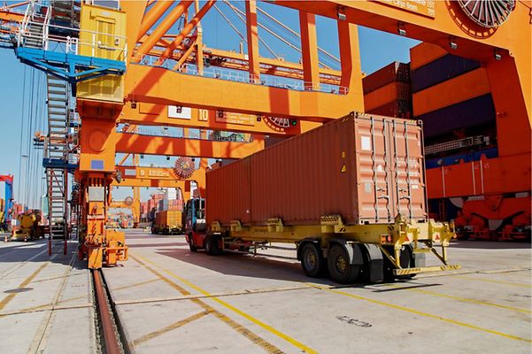 Truck with container at a port beneath large orange loading cranes.