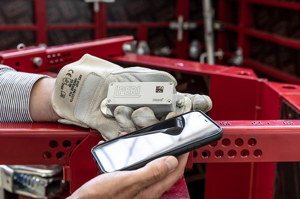 A gloved hand holds a silver PERI sensor next to a smartphone.