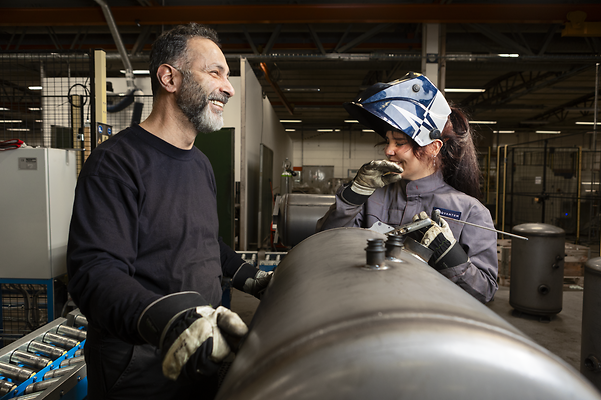Two employees in a production hall are working together on a metal component and chatting to each other with a smile.