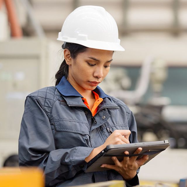 Frau mit Helm in Fabrikhalle schreibt auf ein Tablet