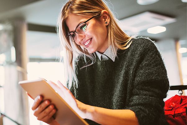 Junge Frau mit Brille sitzt entspannt im Café und arbeitet lächelnd auf einem Tablet.