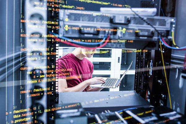 Eine Person arbeitet an einem Laptop in einem Serverraum, umgeben von Serverracks und Netzwerktechnik.