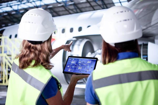 Zwei Frauen mit Sicherheitskleidung und Tablet zeigen auf ein Flugzeug in einer Halle