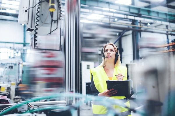 Woman with safety vest and headset in production hall holding clipboard.