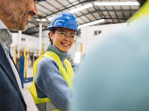 Woman and man standing in an industrial hall, woman smiling and wearing safety helmet